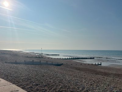 Sunny pebbled beach with wooden groynes stretching into calm blue sea under a clear sky.