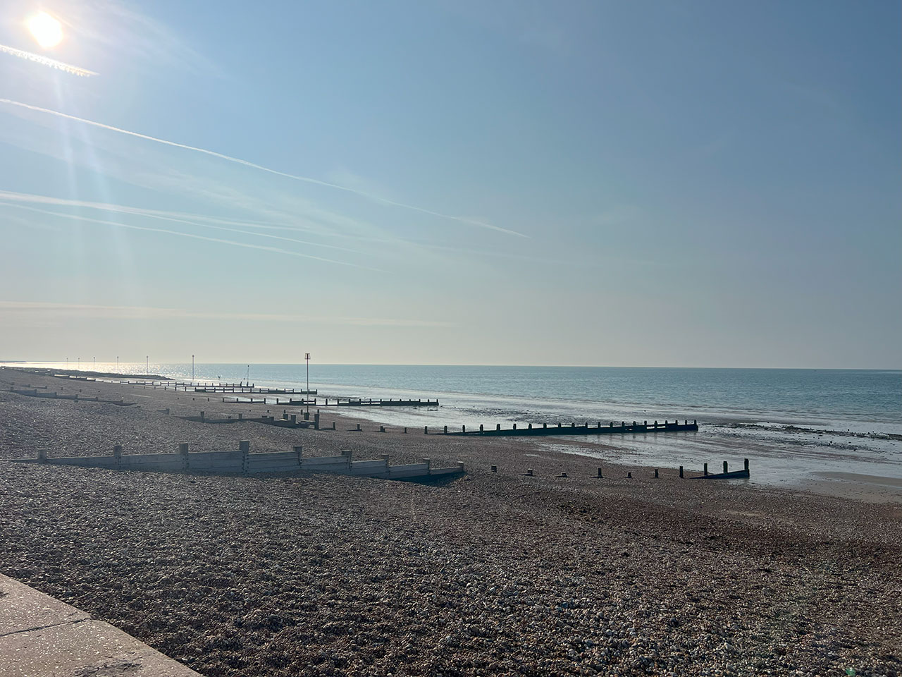 Sunny pebbled beach with wooden groynes stretching into calm blue sea under a clear sky.