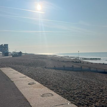 A sunny, clear day on a quiet pebble beach with buildings and the sea in the background.