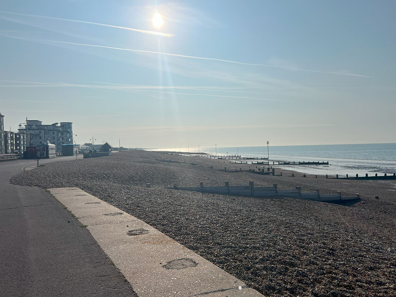 A sunny, clear day on a quiet pebble beach with buildings and the sea in the background.