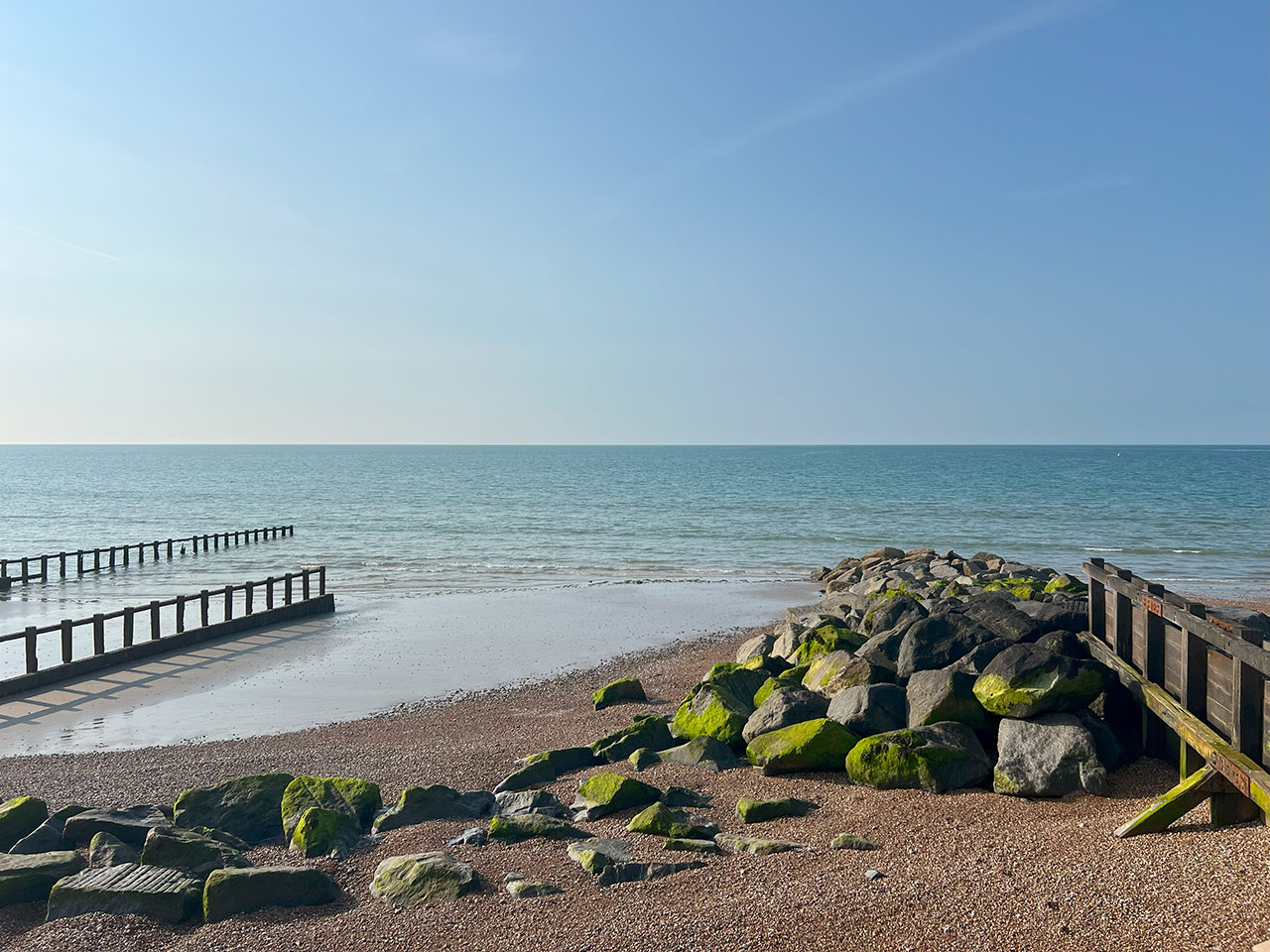 Sandy beach with mossy rocks and wooden groynes, calm sea, and clear blue sky on a sunny day.