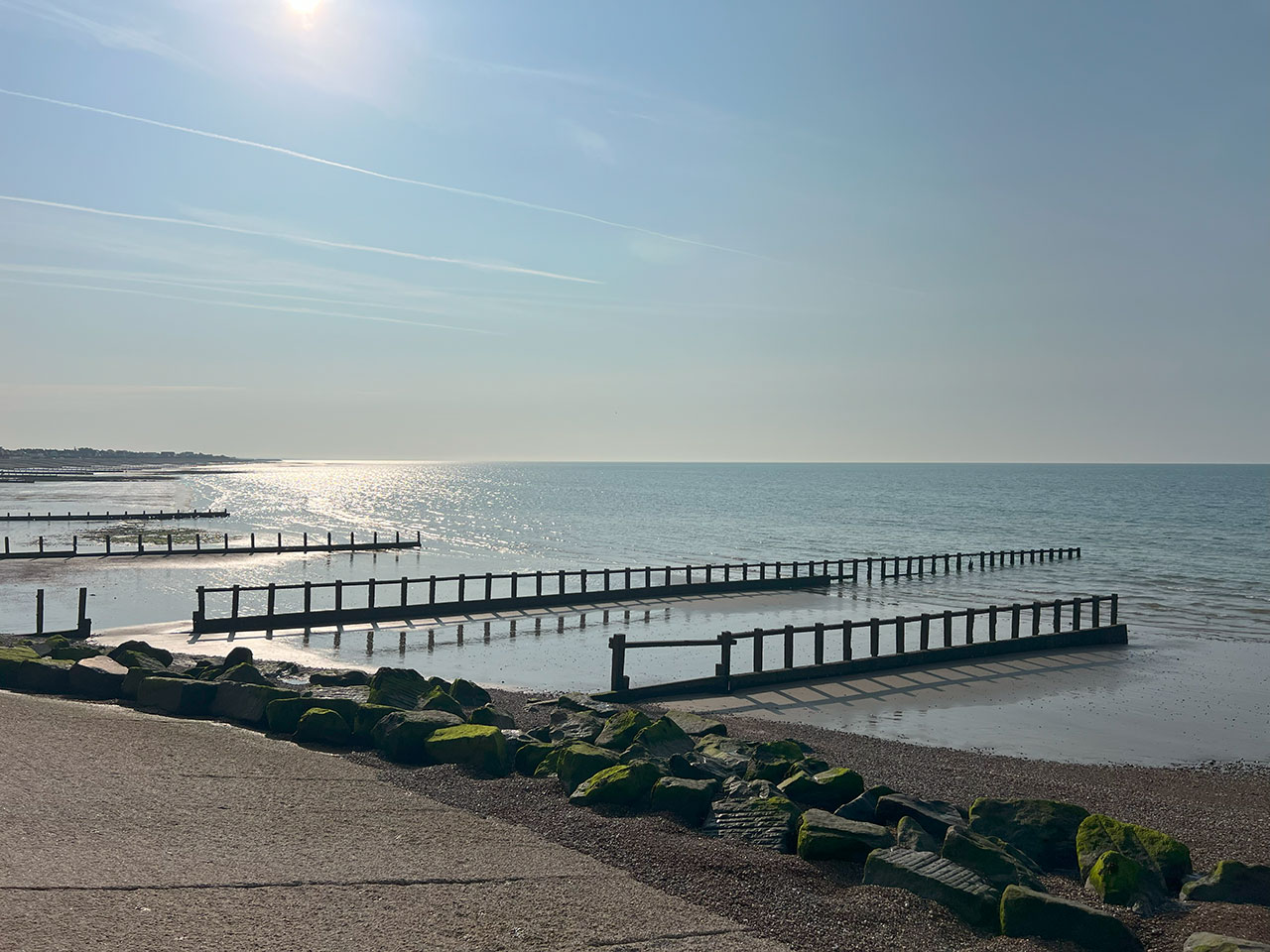 Calm sea view with wooden groynes, rocks with green moss, and sunlight reflecting on the water.