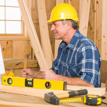 Man in plaid shirt and yellow hard hat building a startup company website, using a hammer and level in a workshop, representing how much a basic website costs.
