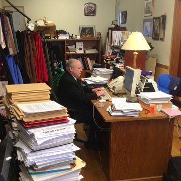 Man working at a cluttered desk with large stacks of papers and folders in a messy office.