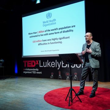 Clive speaks at a TEDx event as a presentation slide displays global disability statistics from the World Health Organization.