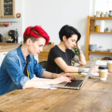 Two teenagers with colourful hair work on laptops at a wooden table in a kitchen.